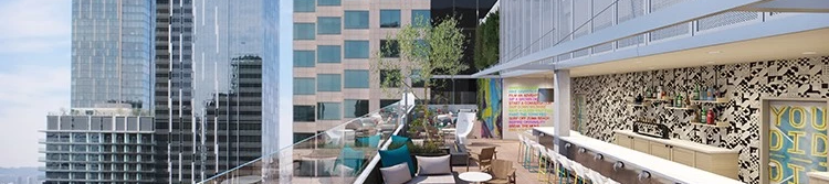 A rooftop terrace with modern chairs and tables, bar area on the right, glass railing, and skyscrapers in the background under a blue sky.
