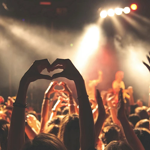 Crowd at a concert, hands raised, stage lights blaze, someone forms a heart shape with fingers—the vibe is energetic and festive. End sentence.