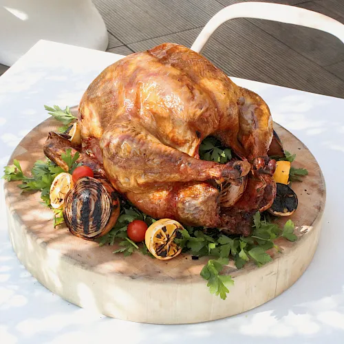 A roasted chicken on a wooden board with herbs, tomatoes, lemons, and garlic, ready to serve.