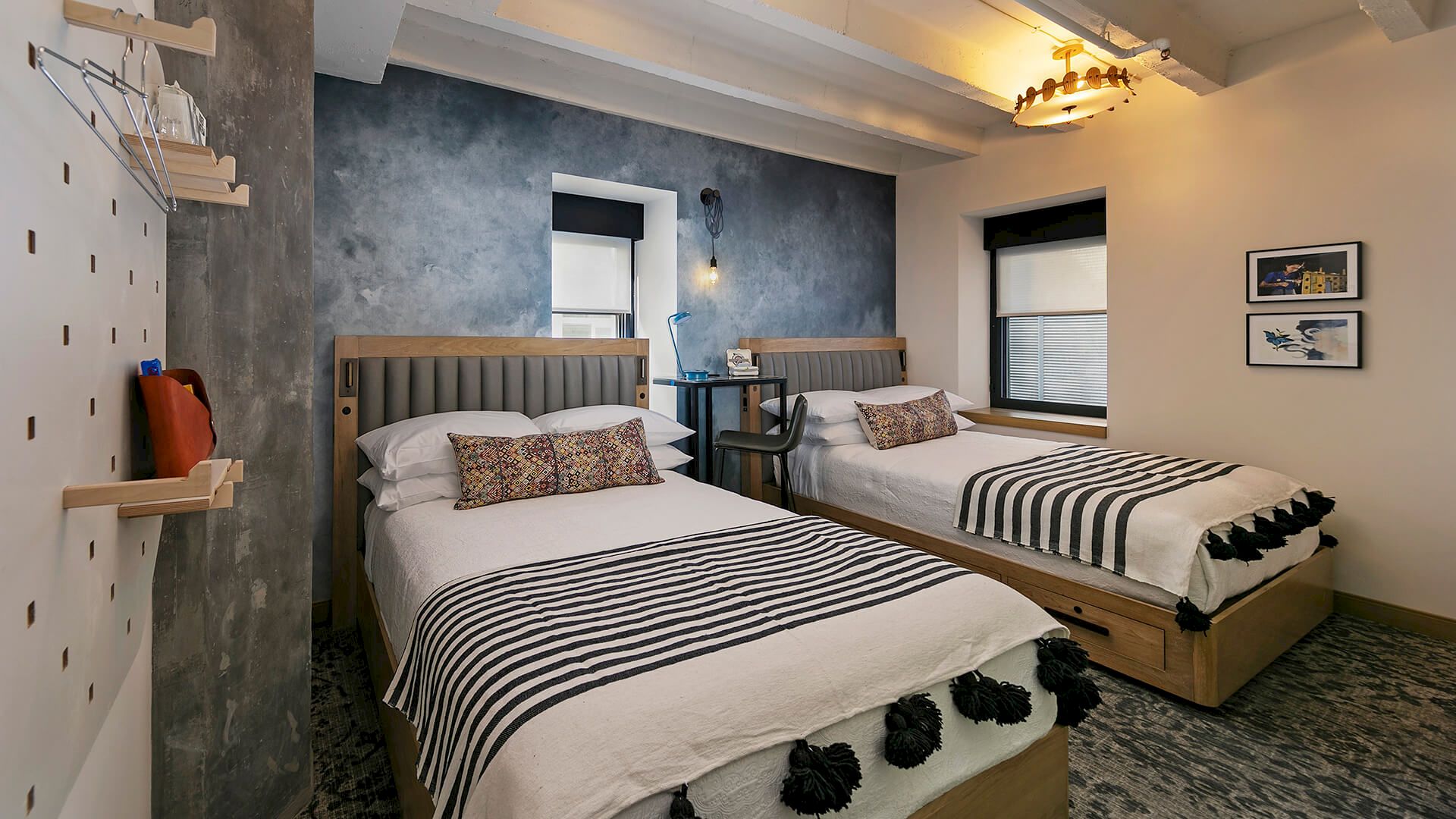A modern twin-bedroom with two beds, a blue textured wall, window blinds, pendant lights, and framed artwork on the walls.
