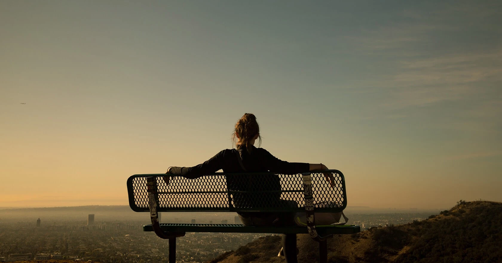 A person sits alone on a bench on a hill at sunset, gazing over a cityscape with warm orange skies and distant buildings.