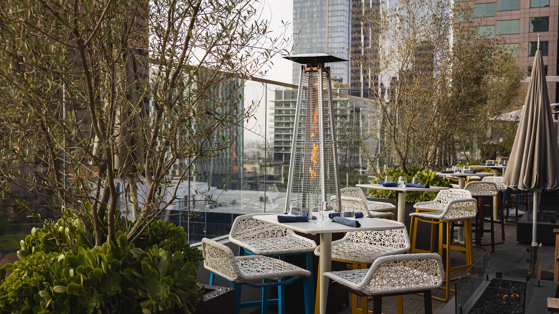 An outdoor restaurant patio with wicker chairs and table, an outdoor gas heater, and greenery beside city office buildings.