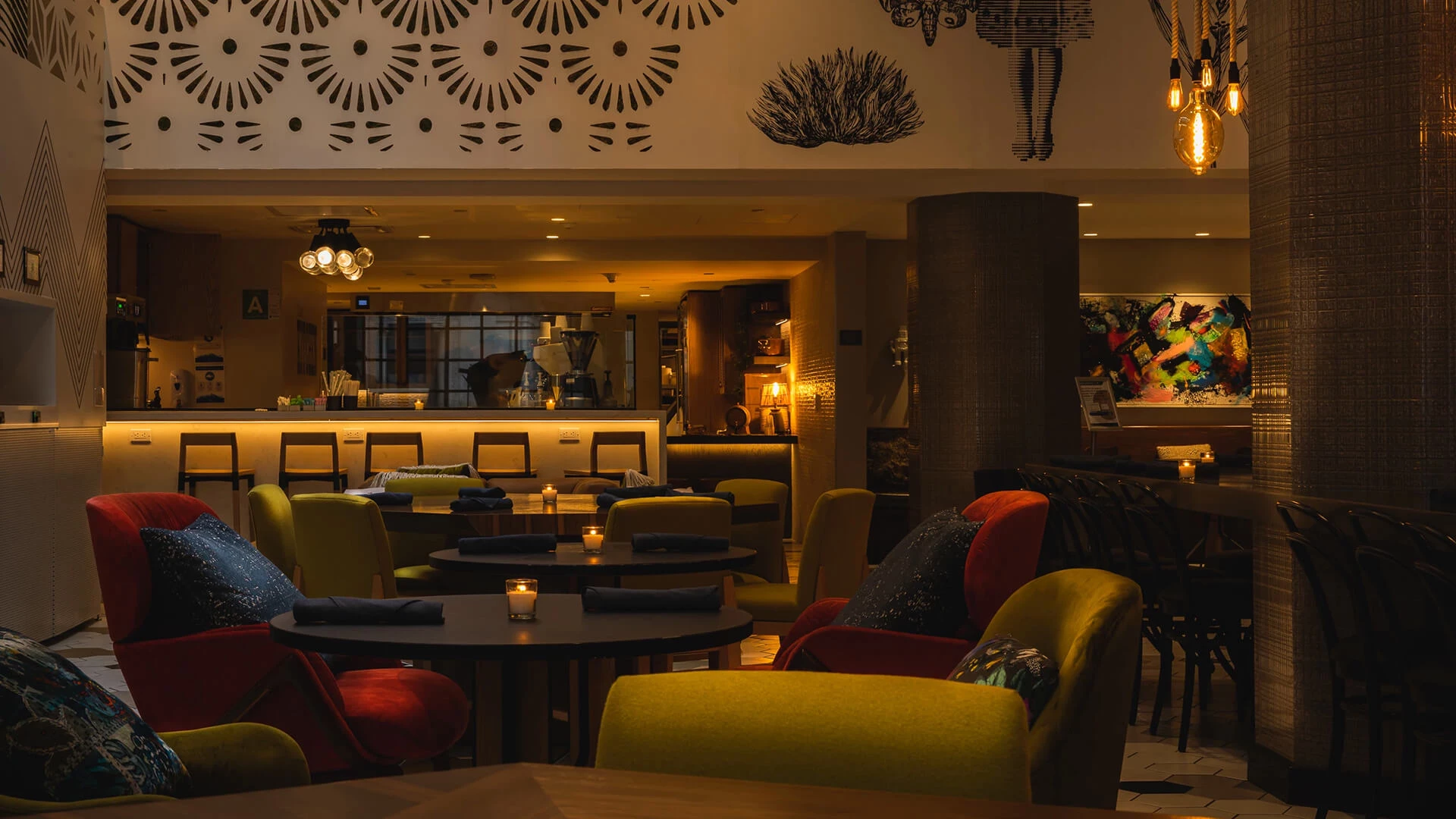 A cozy lounge bar with colorful couches, round tables, warm pendant lights, and a coffee counter in the background.
