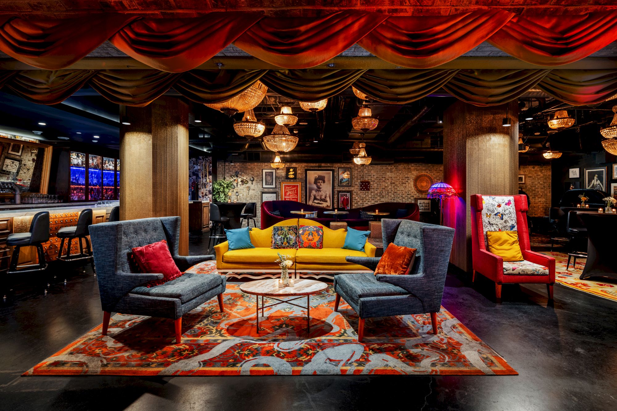 A stylish, colorful lounge with plush blue and yellow seating, vibrant rugs, warm lighting, and art-filled walls in a chic social space.