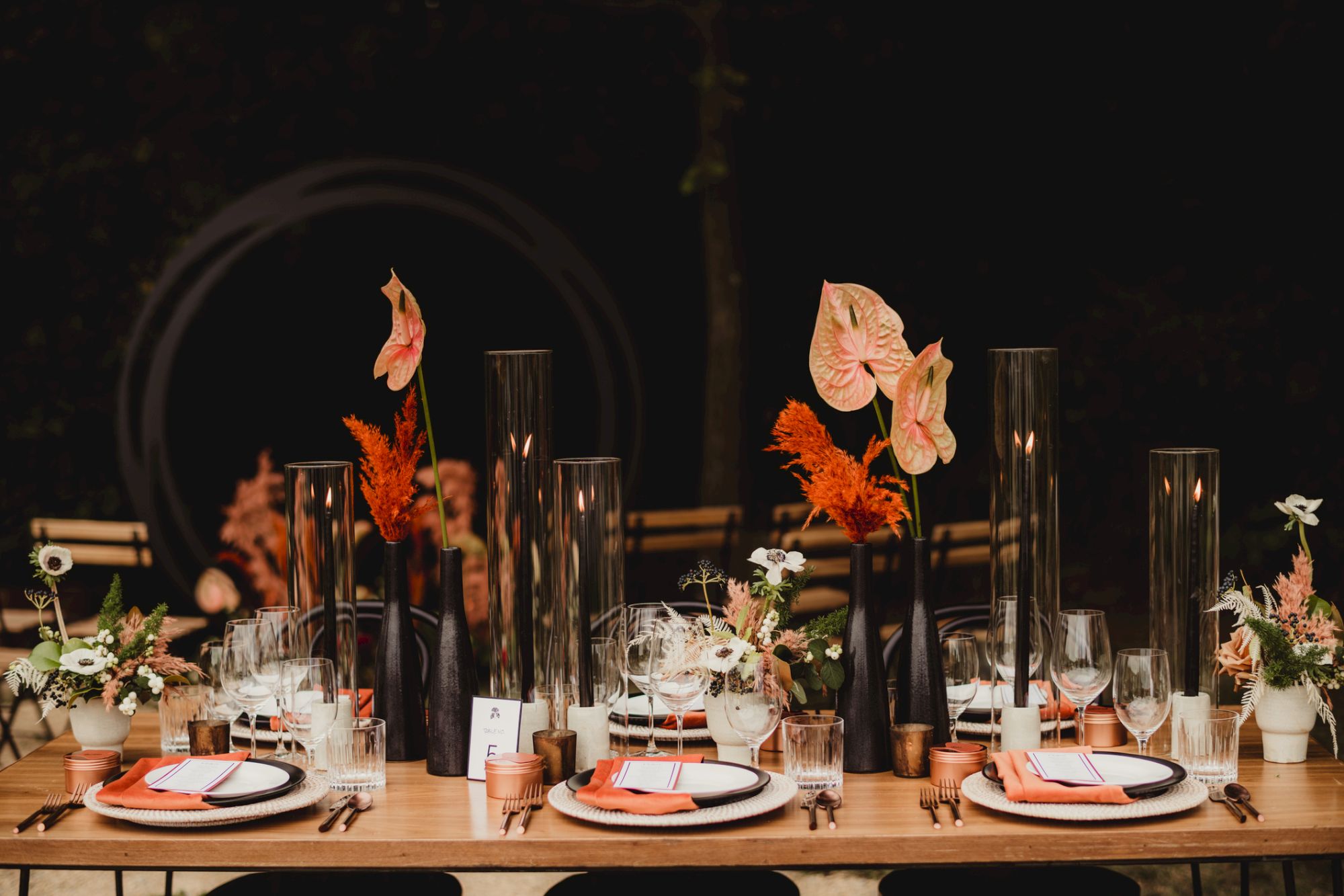 A stylish dinner table setup with white and peach plates, glassware, copper accents, tall black candles, and floral centerpieces in warm autumn tones, nicely arranged.