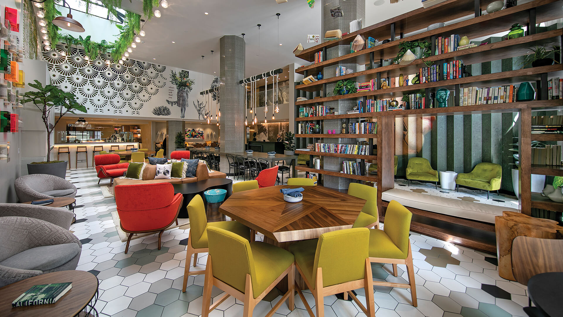 A modern lounge-library with colorful chairs, a large table, bookshelves, greenery, and pendant lights under a decorative mural.