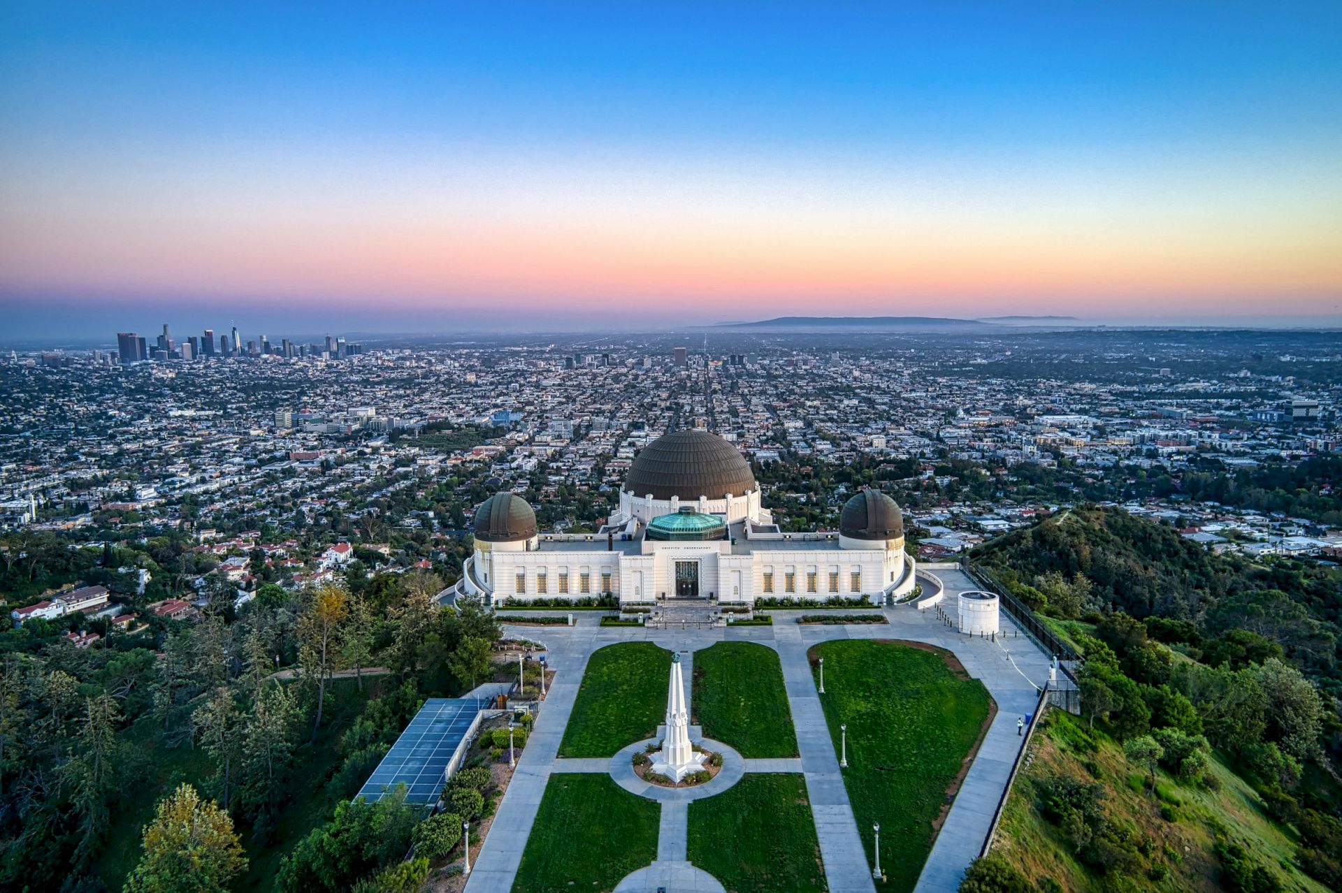 Aerial view of a city skyline at dusk with a large domed observatory and manicured gardens in the foreground.