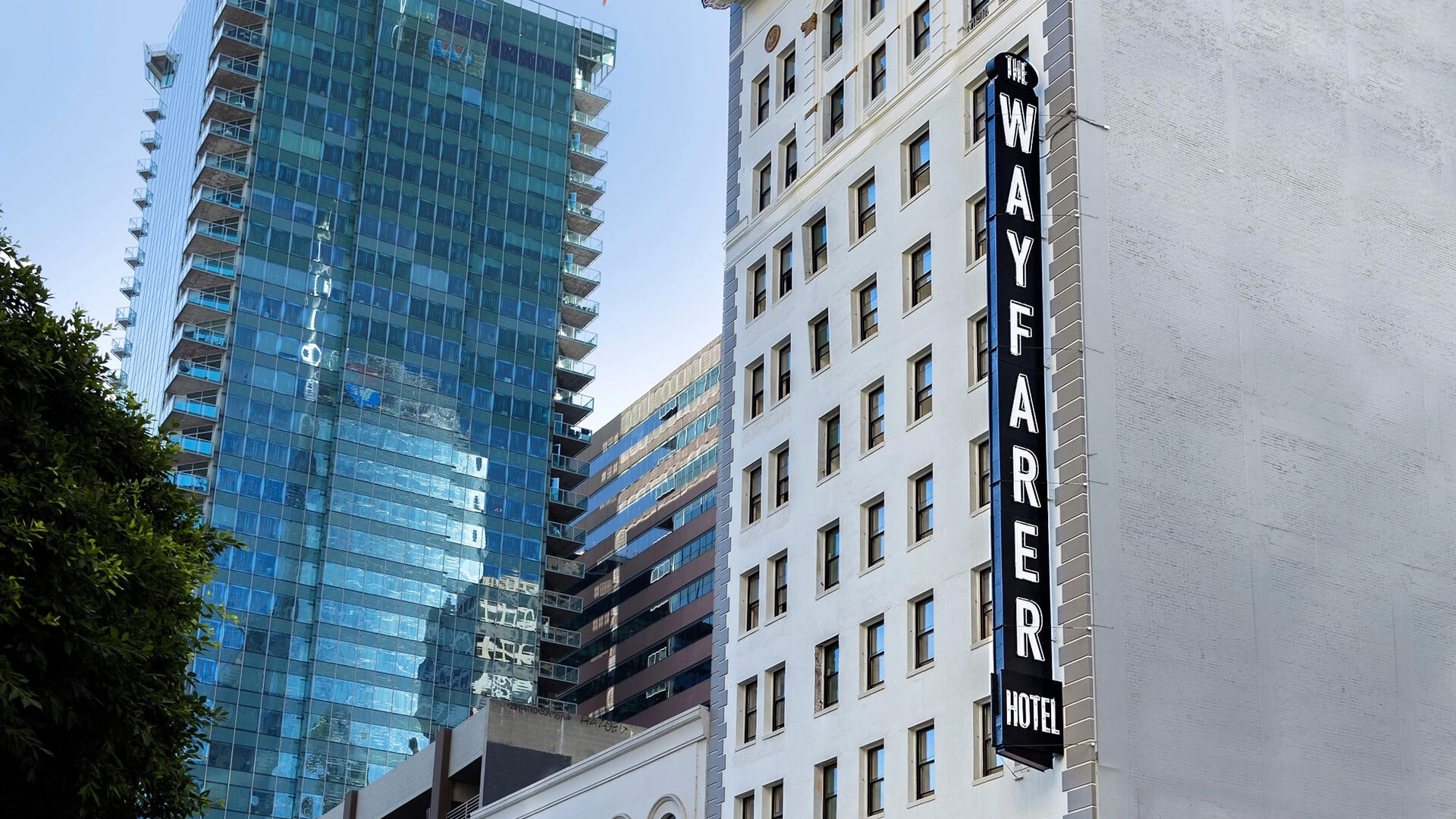 A city street with tall buildings, including a white hotel with a vertical "WAYFARER" sign, and a modern glass-highrise to the left, under a clear sky.