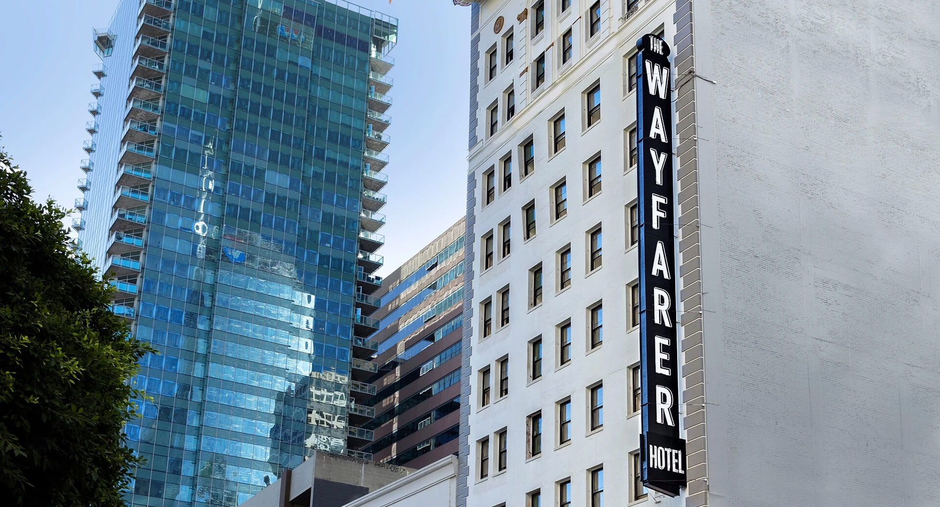 A city street with tall buildings, including a white hotel with a vertical "WAYFARER" sign, and a modern glass-highrise to the left, under a clear sky.