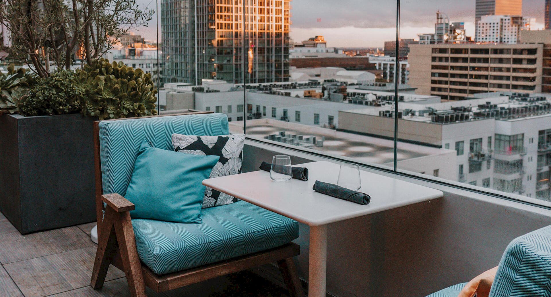 A cozy balcony with turquoise cushions, a small table, and a city skyline backdrop at sunset, perfect for relaxing, ending here.