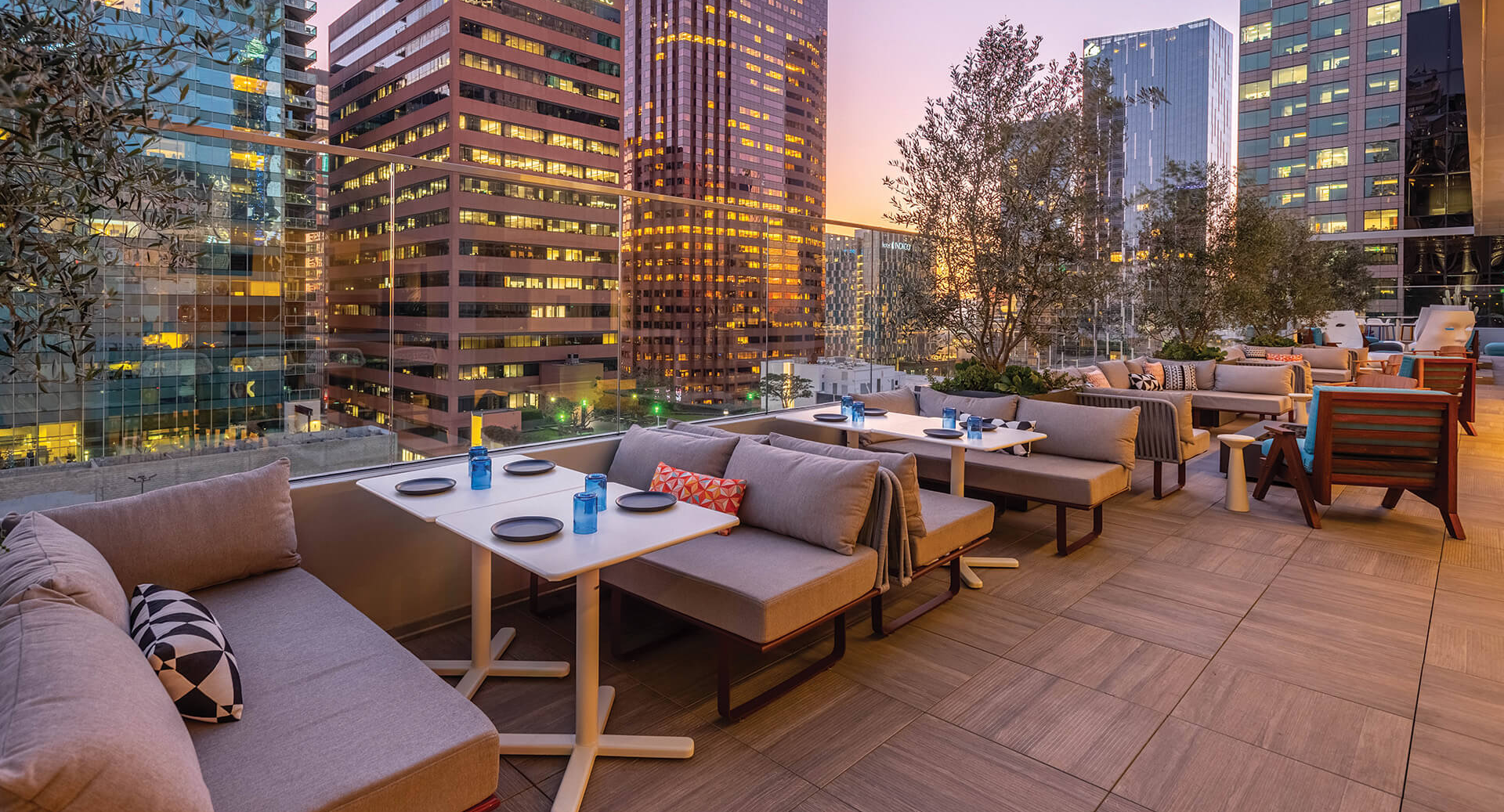A stylish rooftop restaurant with plush seating, white tables, blue glassware, and city skyline views at sunset, perfect for lounging and dining.