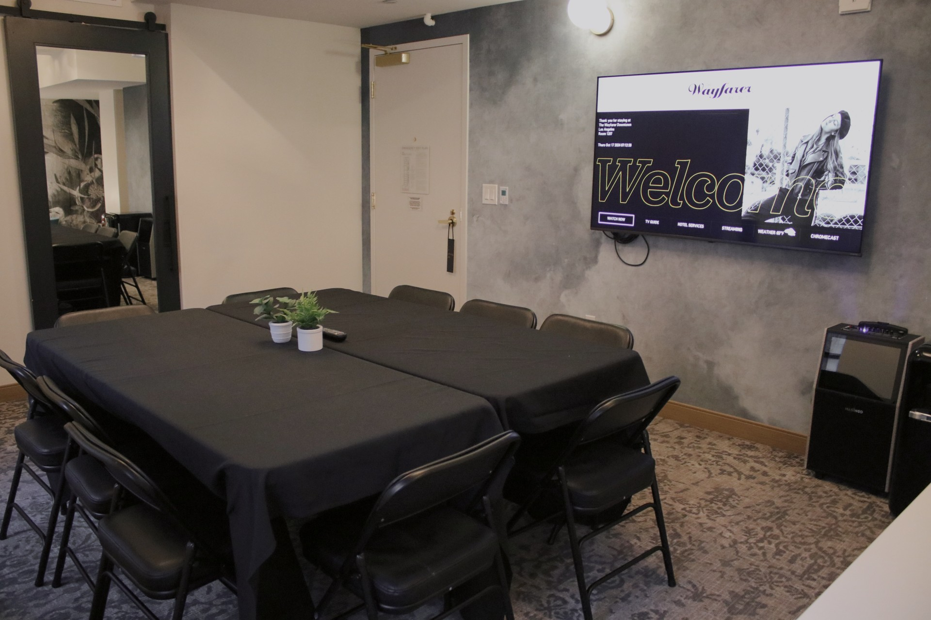 A conference room with a long table, black chairs, a wall-mounted TV, and a small plant centerpiece. 140 characters ending with a period.