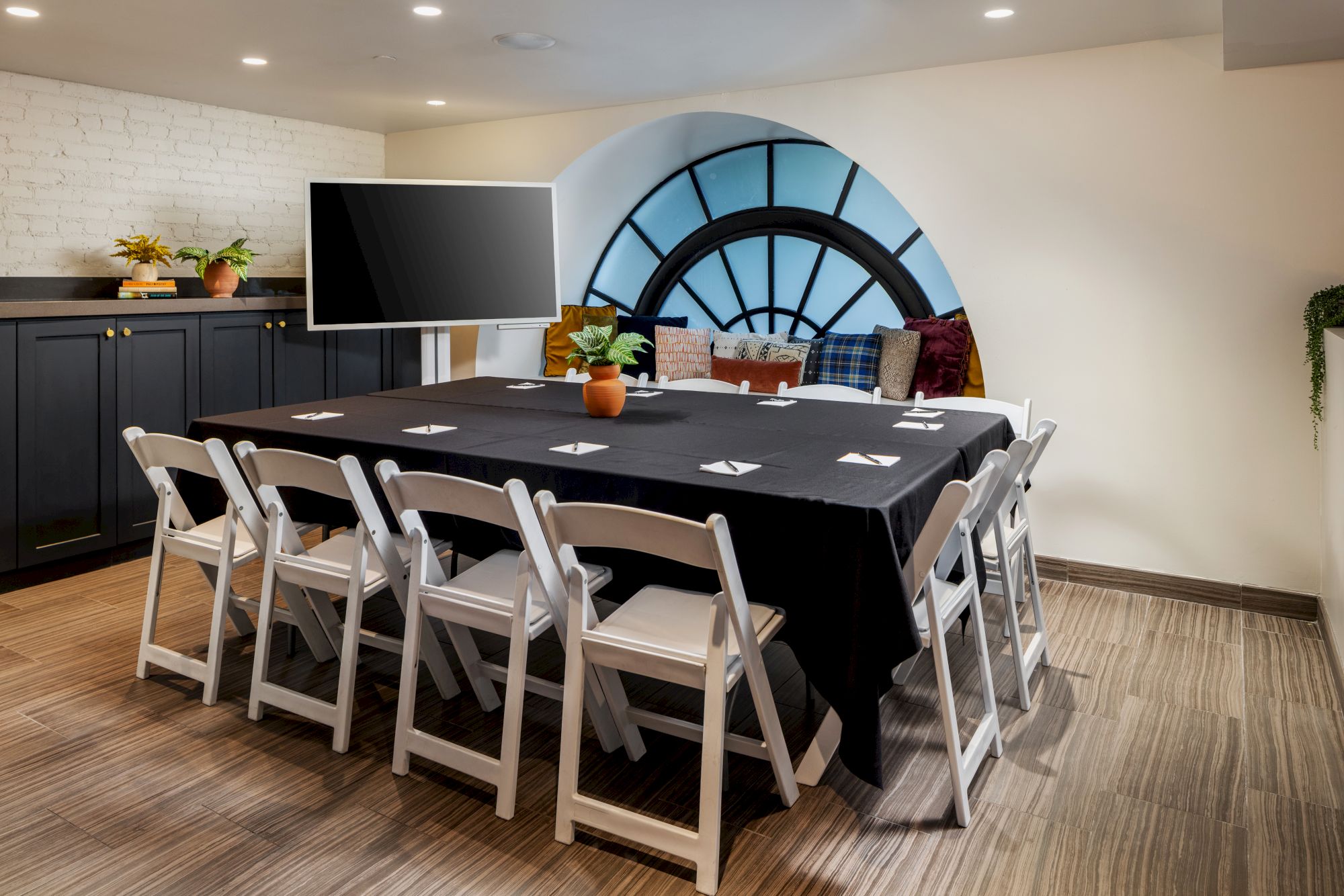 A conference room with a long table covered in a black tablecloth, white folding chairs, a curved window nook, and a TV screen on a sideboard.