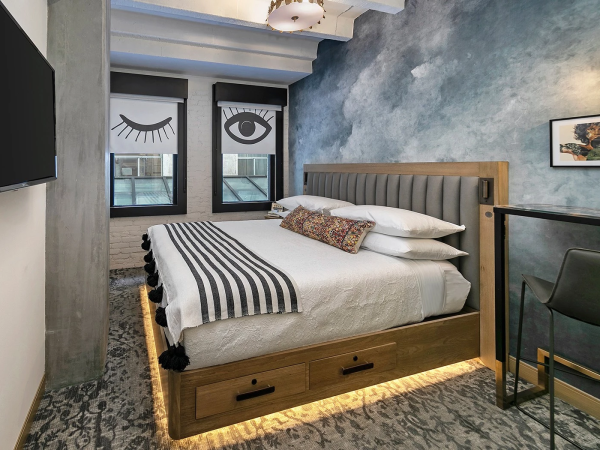 A chic bedroom with a wooden platform bed, built-in storage, striped bedding, and bold eye artwork by the windows; moody blue wall texture.