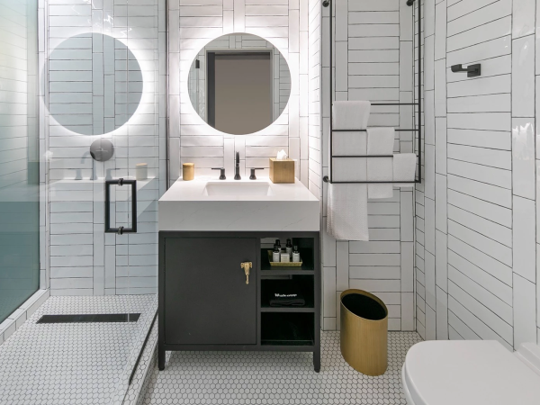 A modern white-tiled bathroom with twin backlit round mirrors, a dark vanity, glass shower with step, towel rack, and a gold trash can.