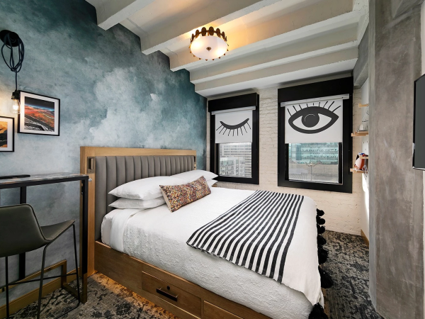 A cozy modern bedroom with a plush bed, striped bedding, and bold window art featuring an eye; a desk with a chair, wall art, and a textured blue-green wall.
