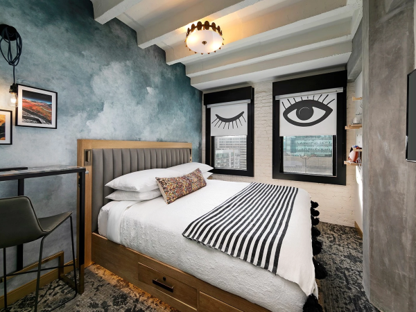 A stylish bedroom with a plush bed, striped duvet, blue textured wall, bold window art featuring an eye, desk area, and a modern ceiling light. End.