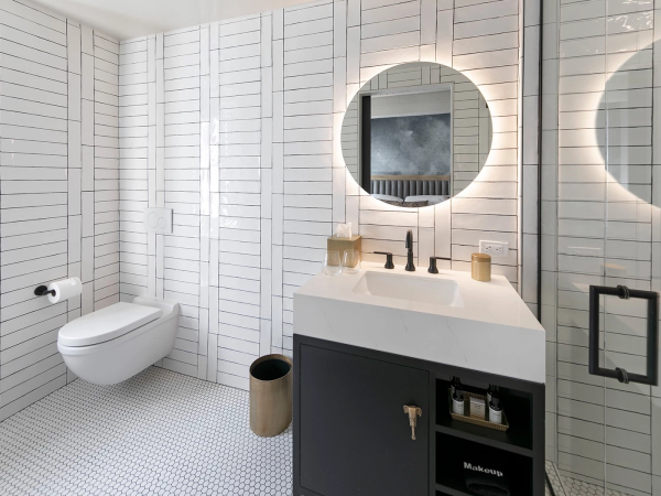 A modern white-tiled bathroom with a circular backlit mirror over a double sink, wall-hung toilet, black vanity, and a frosted glass shower door, minimal and sleek.