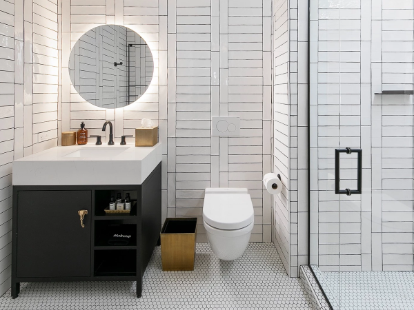 A modern bathroom with white tiled walls, circular mirrors, a dark vanity, sink, stacked storage, wall-mounted toilet, a gold trash can, and a glass shower enclosure.