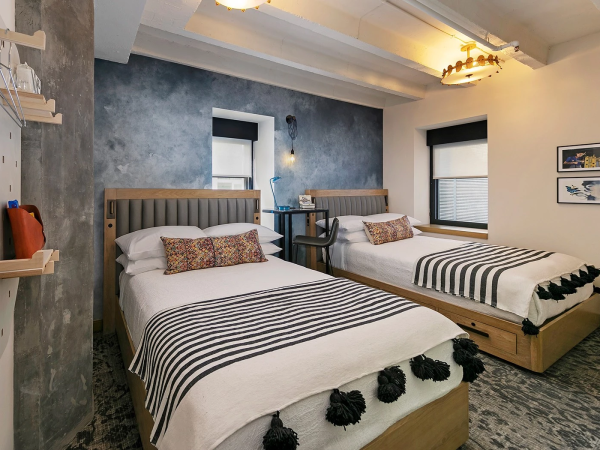 Two twin beds with striped bedding and pom-pom throws, blue accent wall, desk, shelves, and framed art in a modern hotel-style room. ends.