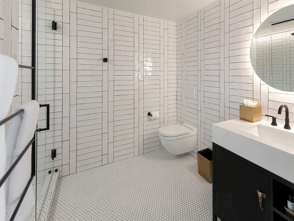 A modern, white tiled bathroom with a wall-mounted toilet, black fixtures, a round mirror, and a dark vanity with a white countertop, towel rack, and tidy decor.