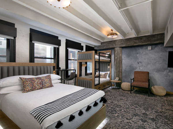 A stylish hotel room with two wooden-frame beds (one queen, one bunk), a modern lounge area, patterned rug, and large windows letting in light.