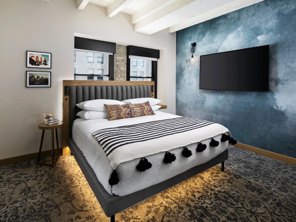 A stylish bedroom with a gray upholstered bed, striped bedding with tassels, a blue textured feature wall, a mounted TV, and warm ambient lighting.