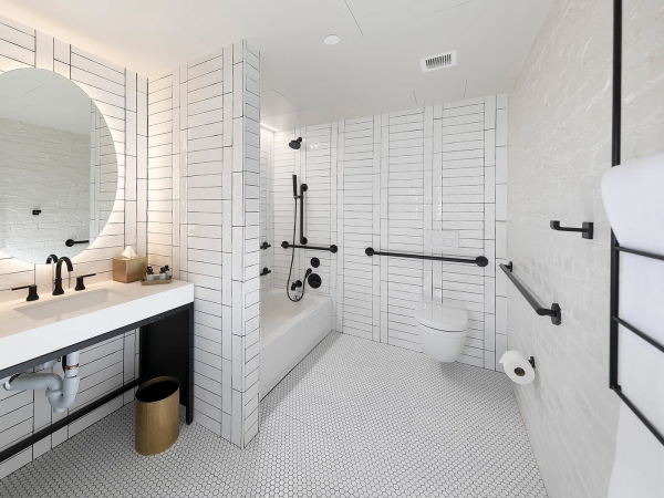 Modern accessible bathroom with white tiled walls, a double sink vanity, round mirror, gold accents, a walk-in shower, grab bars, a wall-mounted toilet, and a black towel rack.