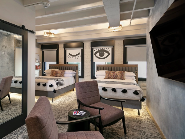 A stylish hotel room with two plush beds, chic seating area, bold eye-wall art, large mirror, and soft lighting on a patterned rug.
