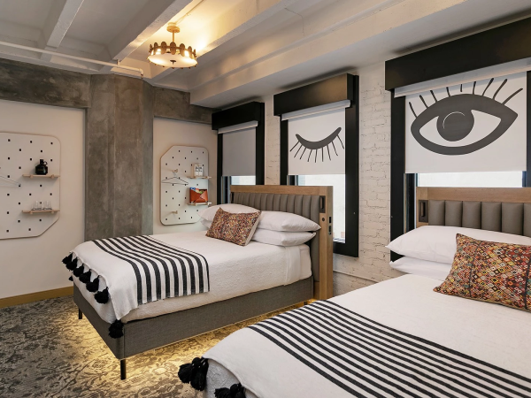 A chic hotel room with two twin beds, striped bedding, bold black-and-white art featuring an eye and a wing motif, and modern decor.