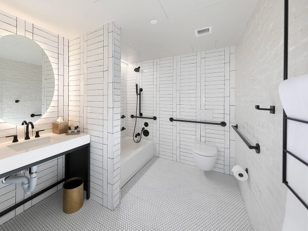 A modern, accessible bathroom with a white subway-tiled shower wall, large round mirror, double vanity, bath, and multiple grab bars for safety.