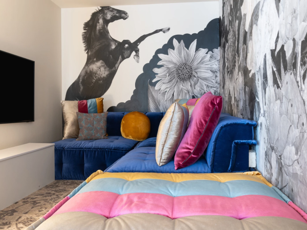 A colorful, cozy living room with a blue sofa, vibrant pillows, a floral mural, and a bright striped bed in the foreground, end of sentence.