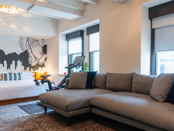A spacious, modern studio with a gray sectional sofa, bed, mural of praying hands, exercise bike, and large windows letting in light.