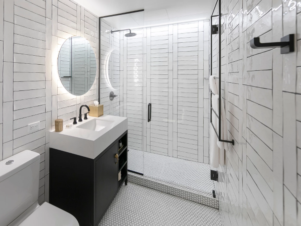 A modern bathroom with a white tiled shower, round backlit mirror, black vanity, small hex tile floor, and neutral accessories, compact and sleek.