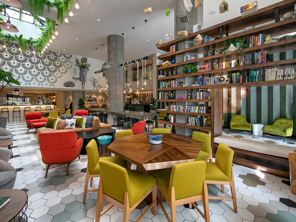 A bright, stylish lounge with colorful chairs around a wooden hexagonal table, bookshelves, greenery, mosaic wall, and cozy seating areas.
