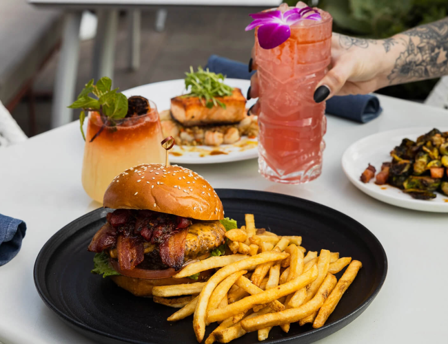 A juicy burger with fries on a black plate, a side of crisp greens, a tall pink cocktail with an orchid, and a colorful vegetable dish in the background.