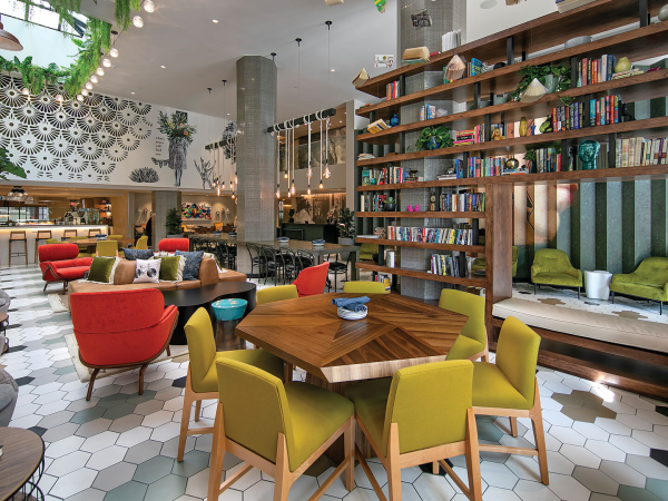A stylish, cozy library-style cafe with colorful chairs, hex tile floor, floor-to-ceiling shelves, plants, and warm ambient lighting. End.