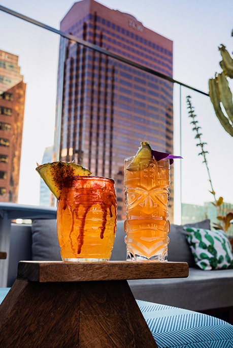 A rooftop lounge scene with cocktails on a wooden serving board, cozy seating, a cactus plant, and skyline skyscrapers in the background.