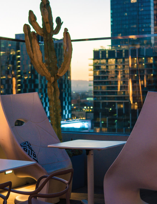 A rooftop seating area at dusk with modern chairs, a tall heater, potted cacti, and abstract mannequin heads near round tables.