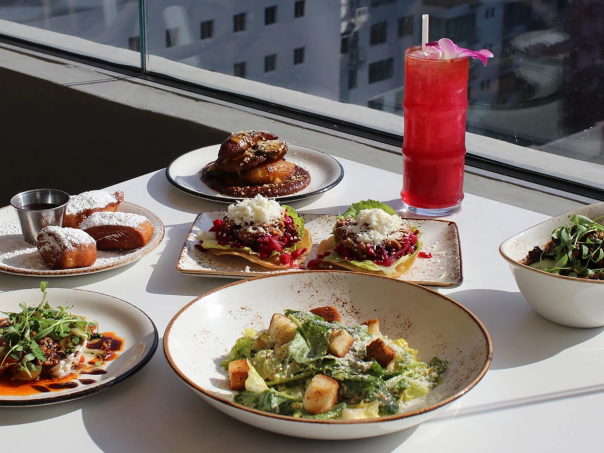A bright rooftop meal: multiple plates of colorful dishes, a creamy greens dish, mini burgers, dumplings, and a tall pink cocktail by a window.