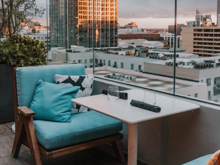 A cozy balcony seating area with blue cushions, a small table, and a city skyline view at sunset.
