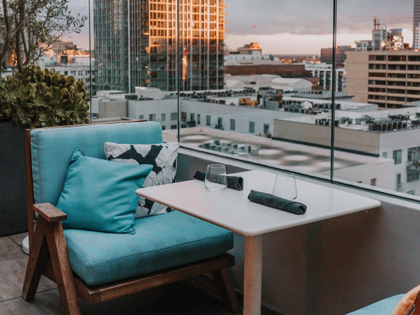A cozy balcony seating area with blue cushions, a small table, and a city skyline view at sunset.