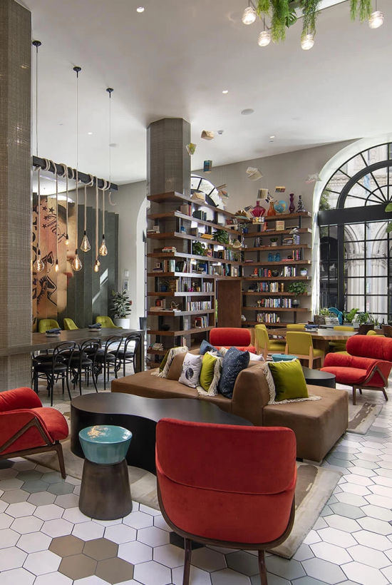 A stylish, cozy cafe with colorful chairs, a long dining table, a mini bar, hanging lights, plants, and a bright arched window in the back.
