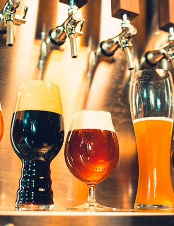 A row of beer glasses with varying colors, from pale to dark, arranged under tall taps in a bar setting.