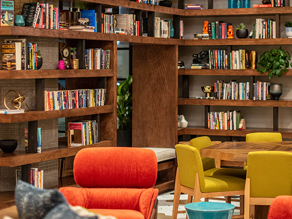 A cozy library lounge with wooden shelves full of books, colorful chairs (orange, yellow), round tables, and plants creating a warm, inviting reading nook.