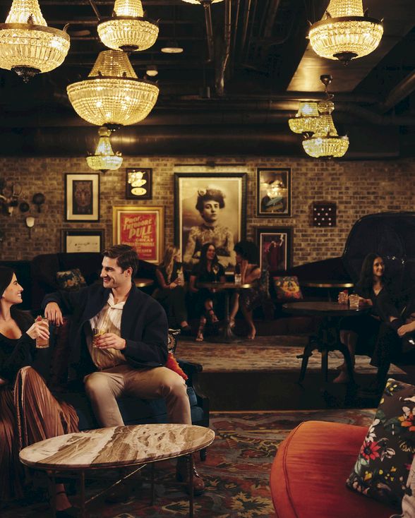 Cozy lounge with friends chatting on couches, warm lighting, mural-art walls, and vintage decor in a dimly lit social space ending with a period.
