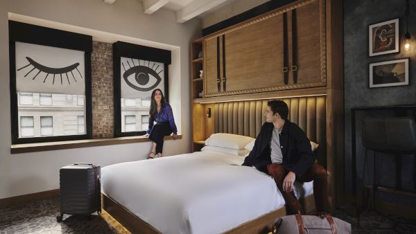 A stylish hotel room with a large bed, two people lounging&mdash;one on the bed&rsquo;s edge and the other near a window displaying bold eye artwork, suitcase nearby.