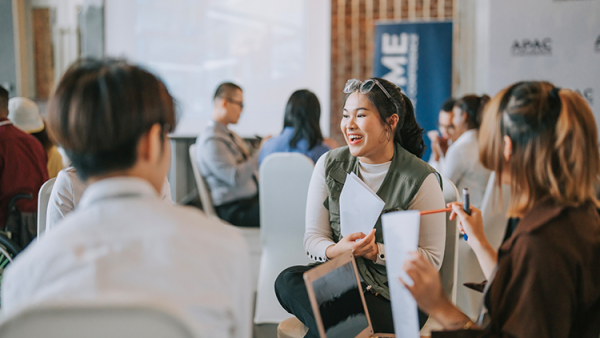 A group of young professionals chat in a bright, modern office or event space, seated in circles, sharing papers and smiles during a discussion.