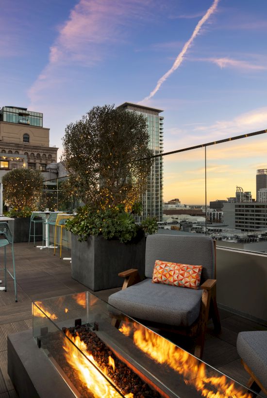 Rooftop lounge with modern outdoor seating, a lit linear fireplace, potted trees, and city skyline at sunset in the background, stylish and inviting.