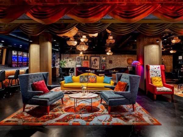 A vibrant lounge with colorful seating&mdash;blue and yellow couches, bright chairs, a patterned rug, warm lighting, and eclectic decor in a cozy, stylish bar.